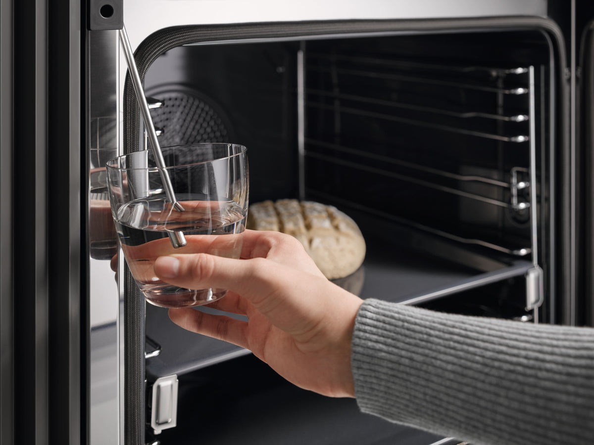 Tuyau d’aspiration plongé dans un verre d’eau devant un four Miele ouvert, pain en cuisson